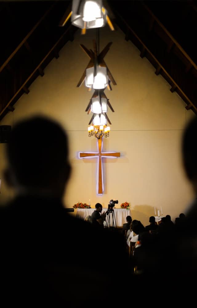 Worshippers facing a cross at the front of a church, with a camera recording the service