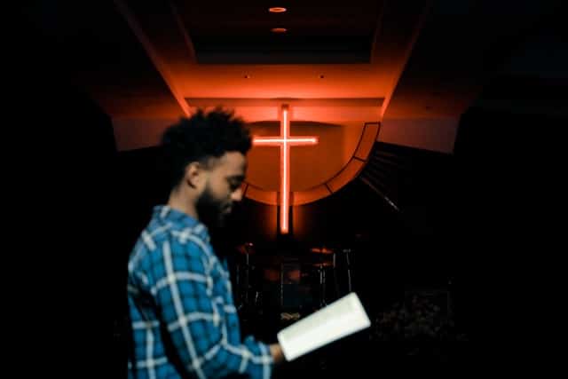 A man holding an open Bible in a church, a glowing cross behind him